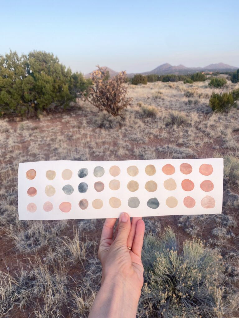 Stella's hand holding a long rectangle of watercolor paper that is covered with painted circles of various earth pigment colors.